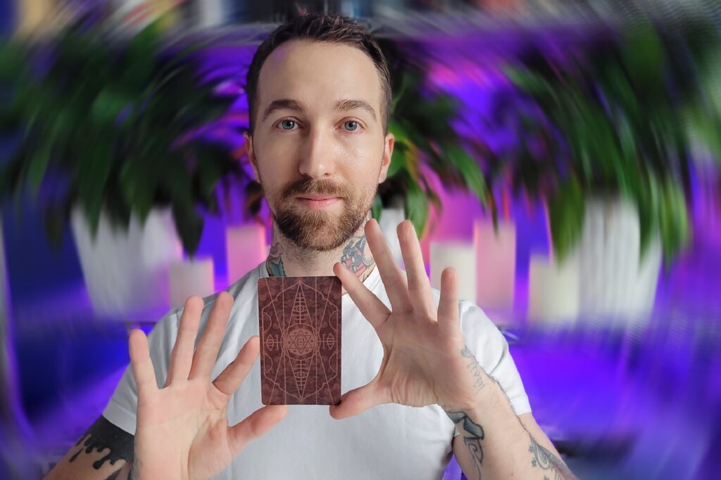 Chris-Bennett-the-best-tarot-reader-near-me-holding-a-tarot-card-from-terra-volatile-with-a-violet-light-background-and-peace-lillies-scaled